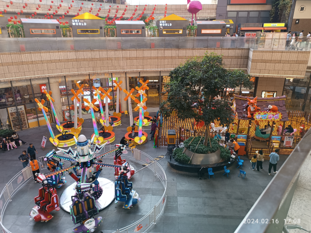 Ein Vergnügungspark innerhalb eines Einkaufszentrums mit Fahrgeschäften, Bäumen, Menschen, Stühlen, Buden und Gebäuden im Hintergrund, mit einem Wasserzeichen in der rechten unteren Ecke.