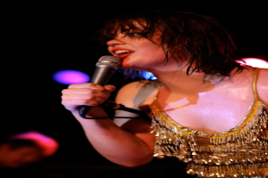 Eine Frau hält ein Mikrofon und spricht auf der Bühne eines Musikkonzerts, mit Lichtern hinter ihr und dem Gesicht eines Mannes in der unteren linken Ecke.