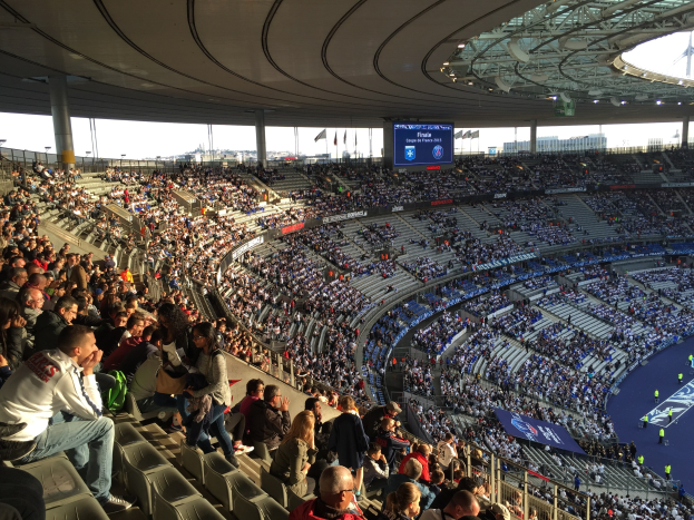 Große Zuschauermenge im Allianz Arena Stadion bei einem Fußballspiel, mit einer Bühne rechts, Fahnen, Stangen und einem Bildschirm im Hintergrund unter einem sichtbaren Himmel.
