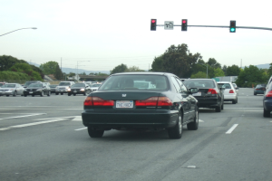 Eine befahrene Straße mit mehreren Fahrzeugen, drei Strommasten und einem Verkehrsschild auf der rechten Seite bei klarem Himmel.