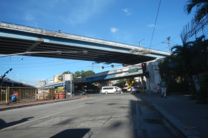 Eine Stadtstraße mit einer Fußgängerbrücke, Fahrzeugen, Fußgängern, Gebäuden und einem bewölkten Himmel.