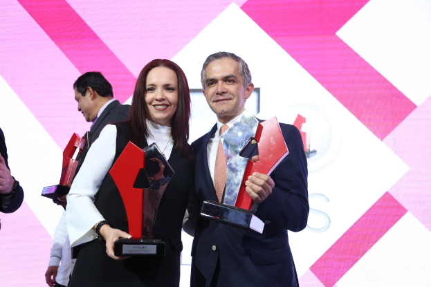 Zwei Menschen, die strahlend und mit Trophäen auf einer Bühne stehen, mit einem Bildschirm im Hintergrund.