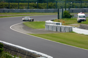 Ein Rennwagen fährt auf einer Rennstrecke mit einem Polizeiwagen im Hintergrund, umgeben von Grünflächen und Absperrungen, mit Text unten.