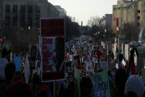 Menschen protestieren auf einer Straße mit Plakaten, mit Gebäuden, Bäumen, Laternenpfählen und dem Himmel im Hintergrund.