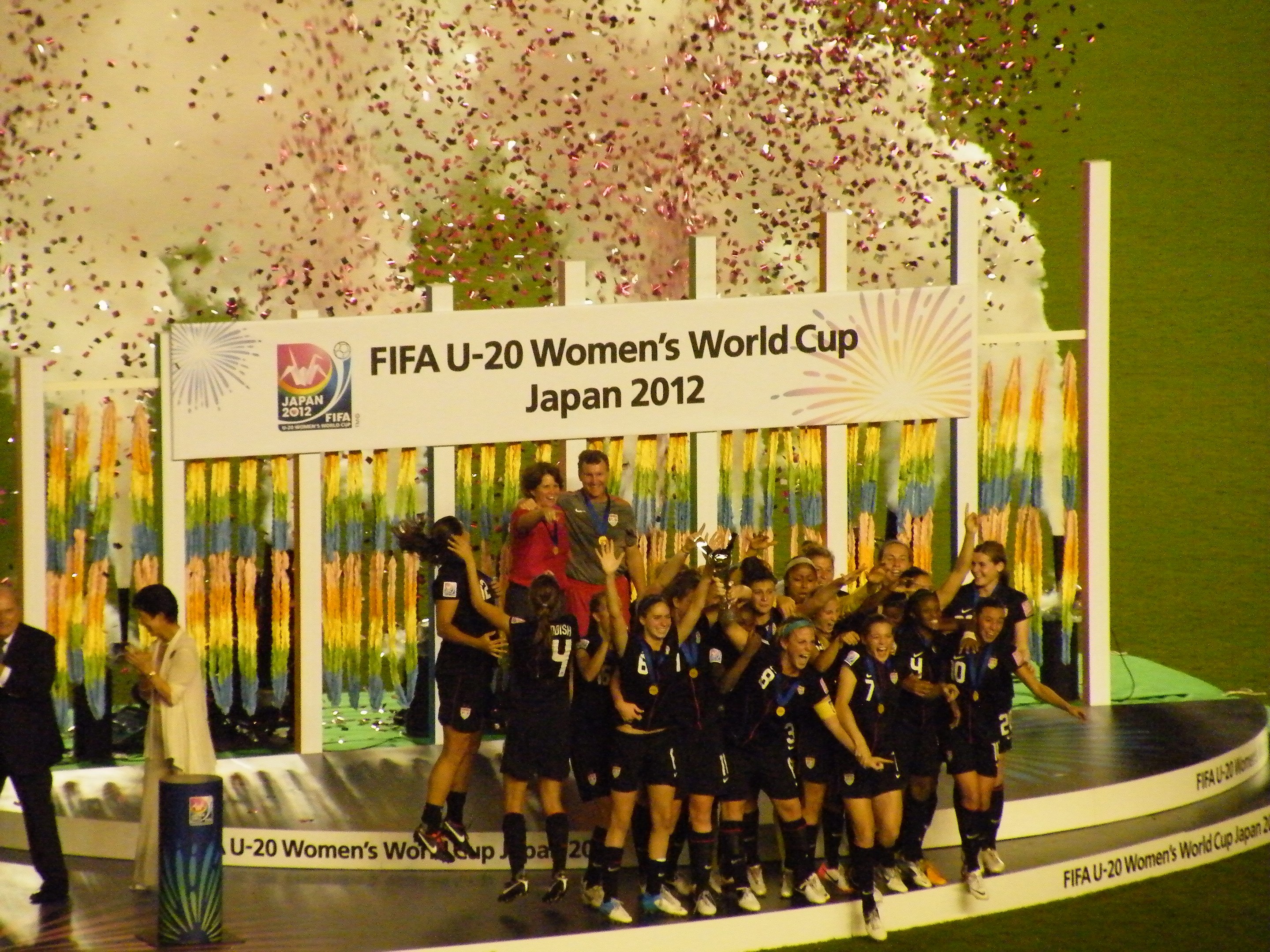 Gruppe von Frauenfußballspielerinnen in schwarzer Uniform, die auf einer Bühne umgeben von einer Menge feiern, mit einem Hintergrundschild und dekorativen Gegenständen auf Gras.
