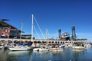 Boote auf dem Wasser im Vordergrund mit einer Brücke, Bögen, Fahnen, Bannern, Gebäuden, Polen, Bäumen und einem bewölkten Himmel im Hintergrund.