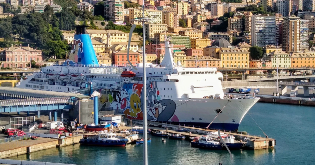 Ein großes Kreuzfahrtschiff liegt nahe einer Stadt mit einer Brücke auf der linken Seite, Fahrzeugen auf der Straße und Gebäuden und Bäumen im Hintergrund.