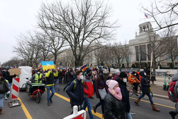 Eine große Gruppe von Menschen marschiert auf einer Demonstration in Washington, D.C. mit Schildern und Bannern, einige fahren Fahrräder, unter einem klaren blauen Himmel.