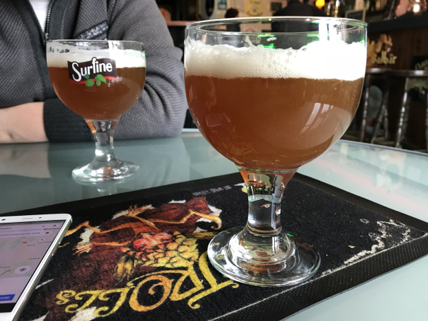 Zwei Gläser goldenes Bier auf einem Tisch neben einem Handy, mit Menschen und gerahmten Wanddekoration im Hintergrund.