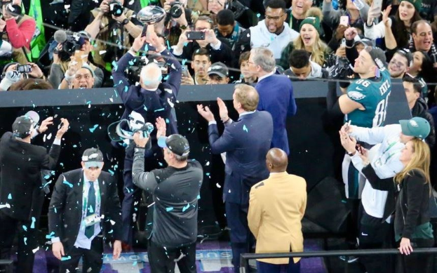 Philadelphia Eagles Spieler feiern mit dem Vince Lombardi Trophy nach dem Sieg im Super Bowl, einige tragen Kappen und halten den Pokal, während andere Konfetti werfen, mit einem Geländer und Fotografen im Hintergrund.