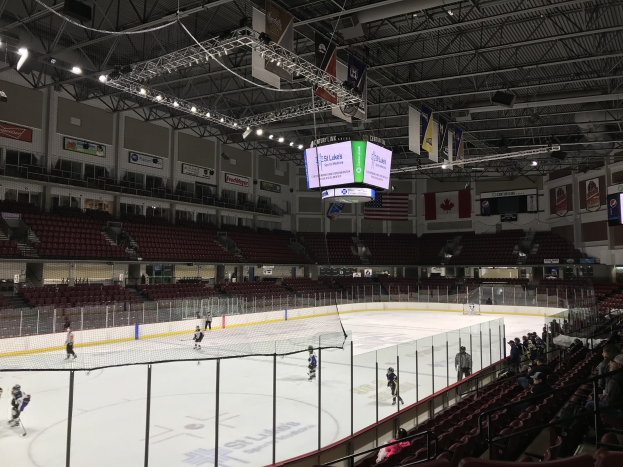 Ein Hockey-Spiel in einer großen Arena mit einem zentralen Netz, Zuschauern auf beiden Seiten, Deckenleuchten und Trägern sowie Bannern an den Wänden, identifiziert als St. Louis Blues Arena.