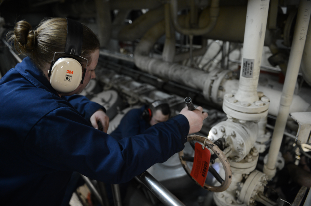 Zwei Frauen in blauen Uniformen arbeiten an einer Maschine in einer Öl- und Gasindustrieumgebung, eine hält ein Objekt und trägt Kopfhörer, mit Rohren und Equipment im Hintergrund.