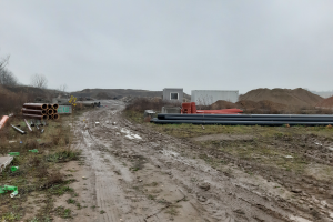 Ein Schotterweg mit zahlreichen Rohren an der Seite, umgeben von Grün, mit einer Hütte und Sand im Hintergrund, unter einem sichtbaren Himmel, wahrscheinlich eine Baustelle für eine Wasseraufbereitungsanlage.