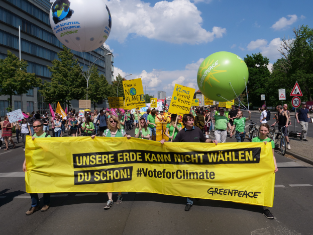 Eine Gruppe von Menschen marschiert mit einer gelben "Wähle für das Klima"-Schleife die Straße entlang, mit Ballons, Schildern, Fahrrädern, Bäumen, Laternenmasten und Gebäuden im Hintergrund unter einem klaren blauen Himmel.