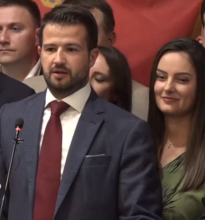 Ein Mann in Anzug und Krawatte steht an einem Rednerpult und spricht in ein Mikrofon, mit einem Banner im Hintergrund, zu einer Menge.