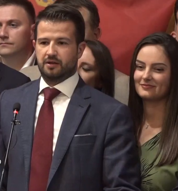 Ein Mann in Anzug und Krawatte steht an einem Rednerpult und spricht in ein Mikrofon, mit einem Banner im Hintergrund, zu einer Menge.