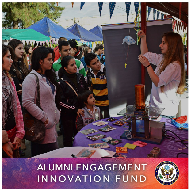 Gruppe von Menschen um einen Tisch versammelt, der ein "Alumni Engagement Innovation Fund"-Schild trägt, mit Gegenständen auf dem Tisch, Zelten und Bäumen im Hintergrund und einem Logo und Text unten.