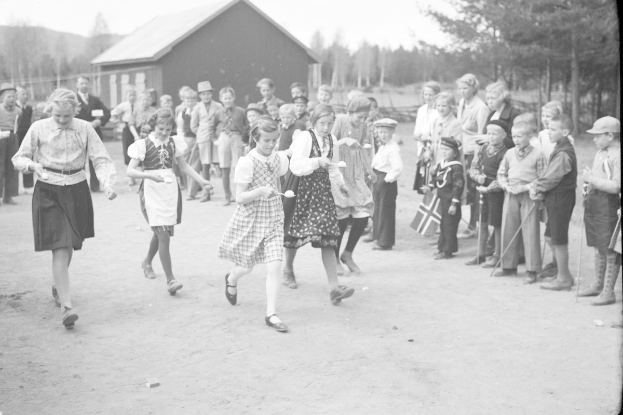 Schwarzes und weißes Foto von Kindern, die vor einer Menge mit Fahnen Fußball spielen, mit Bäumen und einem Haus im Hintergrund.