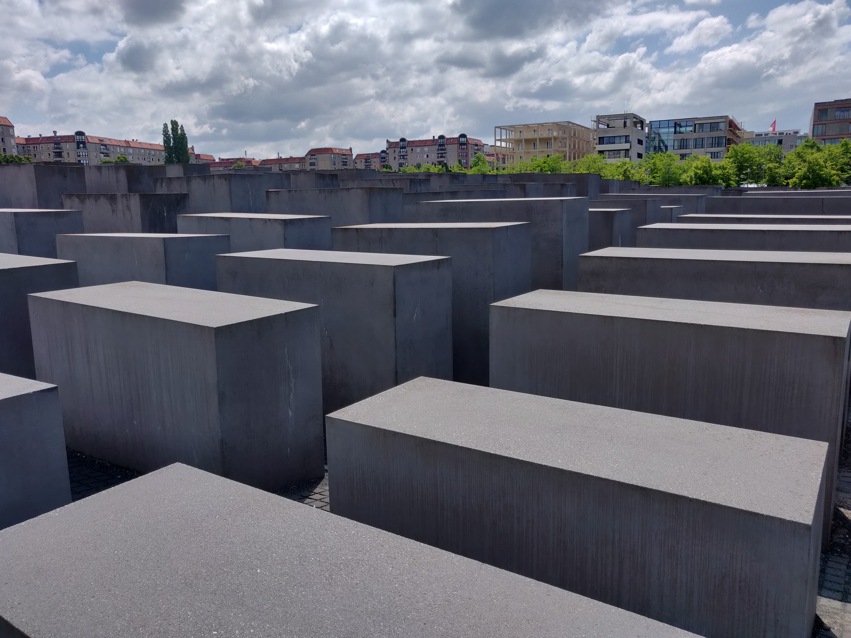 Betonsockel in einem Gittermuster am Holocaust-Mahnmal für die ermordeten Juden Europas in Berlin, mit Bäumen, Gebäuden und Wolken im Hintergrund.