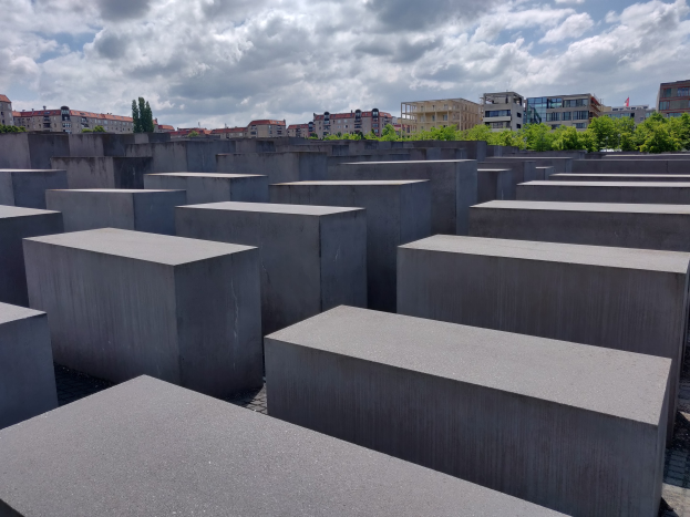 Betonsockel in einem Gittermuster am Holocaust-Mahnmal für die ermordeten Juden Europas in Berlin, mit Bäumen, Gebäuden und Wolken im Hintergrund.