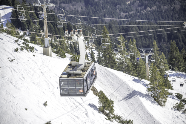 Eine mit Menschen gefüllte Seilbahn fährt einen schneebedeckten Berg hinauf, mit hohen Bäumen im Hintergrund.