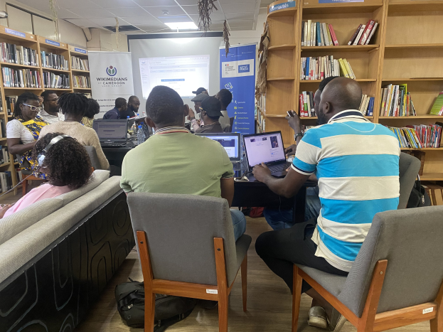 Gruppe von Menschen an einem Tisch mit Laptops umgeben von Bücherregalen, mit einem Banner, Bildschirm, Deckenbeleuchtung und Taschen auf dem Boden im Hintergrund.
