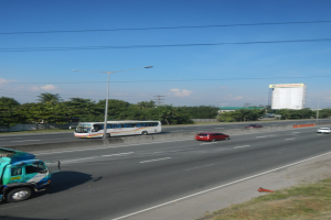 Ein Lkw fährt auf einer Autobahn neben einem Bus, umgeben von Gras, Verkehrskegeln, Straßenlaternen, Strommasten mit Drähten, Bäumen, Gebäuden und einem bewölkten Himmel.