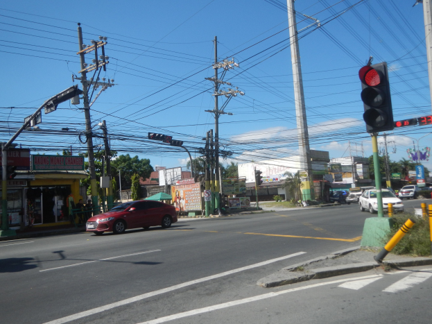 Ein rotes Auto fährt an einer Ampel auf einer Straße vorbei, die von Strommasten und -kabeln gesäumt ist, mit Gebäuden, Bäumen und einem klaren blauen Himmel im Hintergrund.