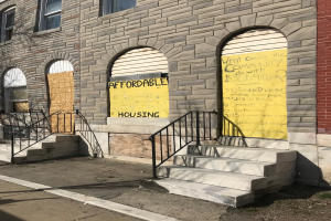 Ein zweistöckiges Gebäude mit vernagelten Fenstern, beschriftet mit "Bezahlbarer Wohnraum", erreichbar über Treppen mit Geländern, neben einer Straße gelegen.