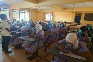 Ein Klassenraum in Nigeria mit Schülerinnen und Schülern, die Gesichtsmasken tragen und auf Bänken sitzen oder stehen, mit Büchern und Gegenständen auf Tischen, Fenstern, einer Tür und Deckenlampen und Ventilatoren im Hintergrund.