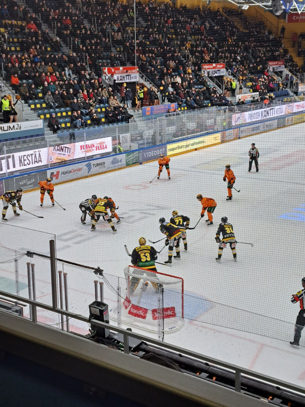 Eine Gruppe von Menschen, die Hockey auf einem Eisring mit einem Netz und einer Umzäunung spielen, Zuschauern im Hintergrund und Bannern mit Text, unter Stadionbeleuchtung.