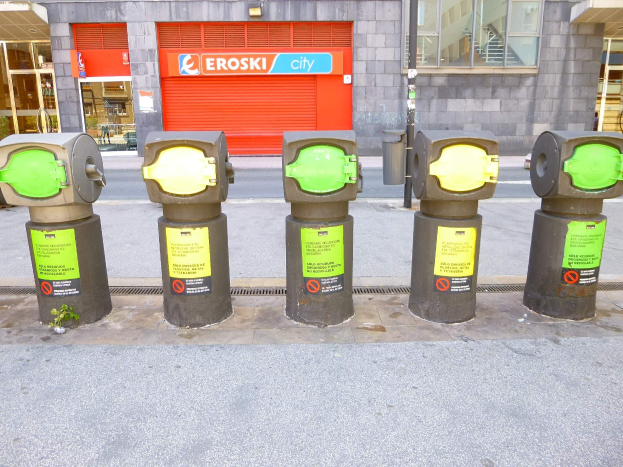 Eine Reihe von Parkautomaten entlang einer Straße mit einem Gebäude, das Glasfenster und eine Rolllade mit Text im Hintergrund und einen nahen Müllcontainer.