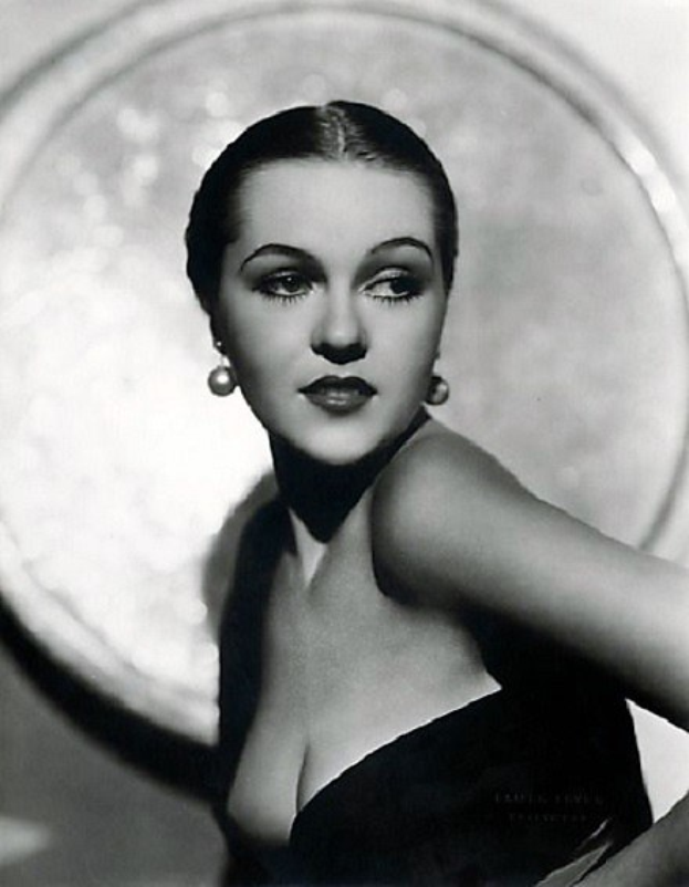 Ein Schwarz-Weiß-Foto einer Frau in einem langen, hochgeschlossenen Kleid mit 1940er-Jahre-Styling und Make-up, die selbstbewusst in die Kamera blickt und vor einem unscharfen Hintergrund und Text am unteren Rand posiert.