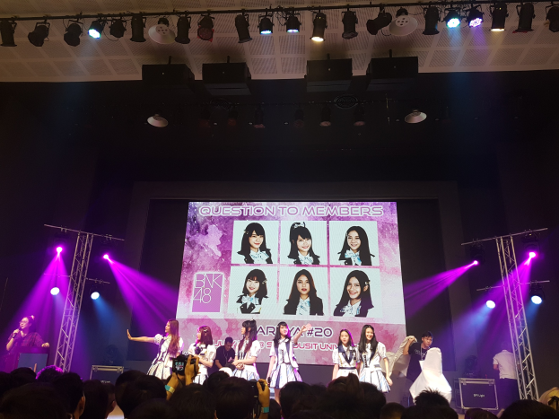 Eine Gruppe von Menschen performs auf einer hell beleuchteten Bühne mit Trägern, während ein Sänger ein Mikrofon hält und ein anderer ein Instrument spielt, während eine Menge mit Kameras unten und ein großer Bildschirm Bilder der K-Pop-Gruppe BK48 im Hintergrund anzeigt.