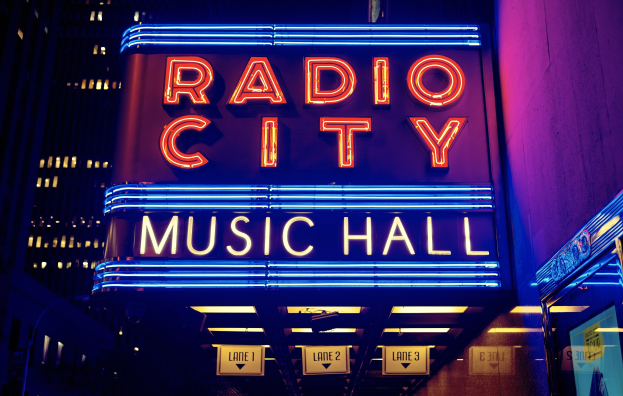 Hell erleuchtetes Radio City Music Hall in New York City mit hohen umliegenden Gebäuden und einem "Radio City"-Schild sowie mehreren kleineren Schildern an seiner Basis.