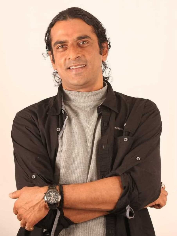 Ein Mann in einem schwarzen und aschgrauen Outfit mit verschr├Ąnkten Armen, der eine Uhr tr├Ągt und gegen einen wei├čen Hintergrund steht und nachdenklich wirkt.