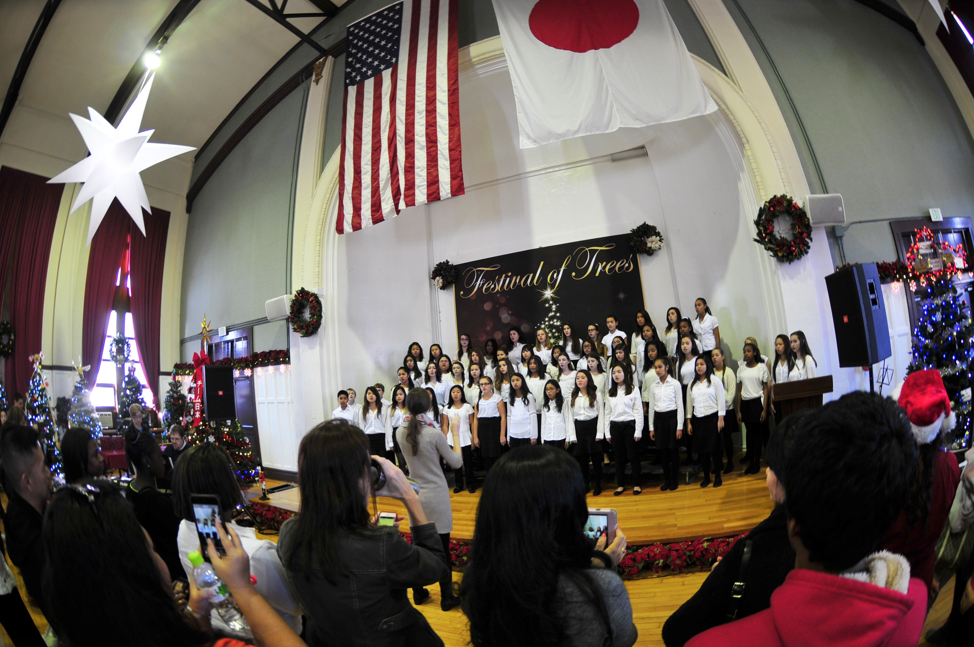 Gruppe von Menschen auf einer Bühne mit Weihnachtsdekoration, einem Rednerpult, Lautsprechern, einer Texttafel und einem Publikum mit Kameras und Handys.