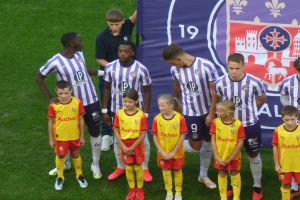 Eine Gruppe junger Menschen auf einem saftig grünen Fußballfeld, mit einem Banner im Hintergrund, auf dem 'Ligue 1' steht, einige tragen Trikots und andere Shorts, unter einem hellblauen Himmel.
