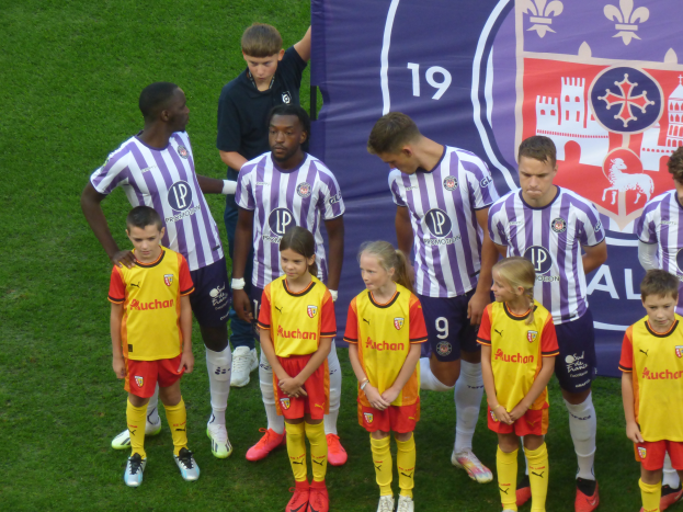 Eine Gruppe junger Menschen auf einem saftig grünen Fußballfeld, mit einem Banner im Hintergrund, auf dem 'Ligue 1' steht, einige tragen Trikots und andere Shorts, unter einem hellblauen Himmel.