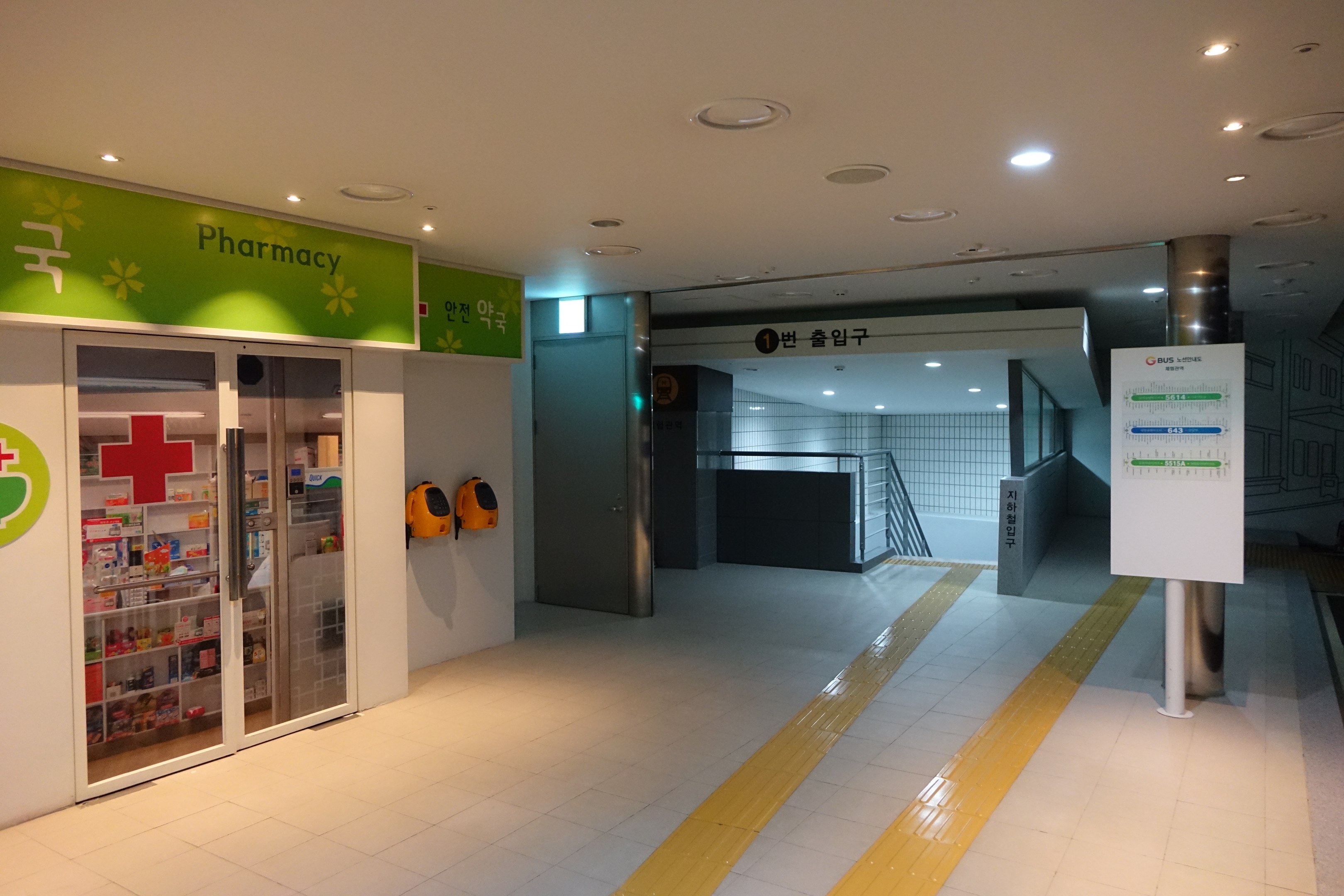 Apothekeneingang mit einem "Apotheke"-Schild auf der linken Seite, einer Tafel mit Text auf der rechten Seite, beleuchteten Lampen auf dem Dach und Gegenständen im Hintergrund.