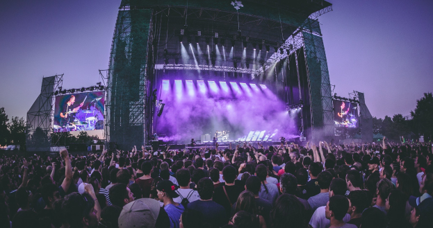 Eine große Menge von Menschen auf einem Musikfestival mit einer Bühne, auf der Musiker Instrumente spielen, beleuchtet von Lichtern und umgeben von Bildschirmen, vor einer Kulisse aus einem Gebäude, Bäumen und einem klaren blauen Himmel.