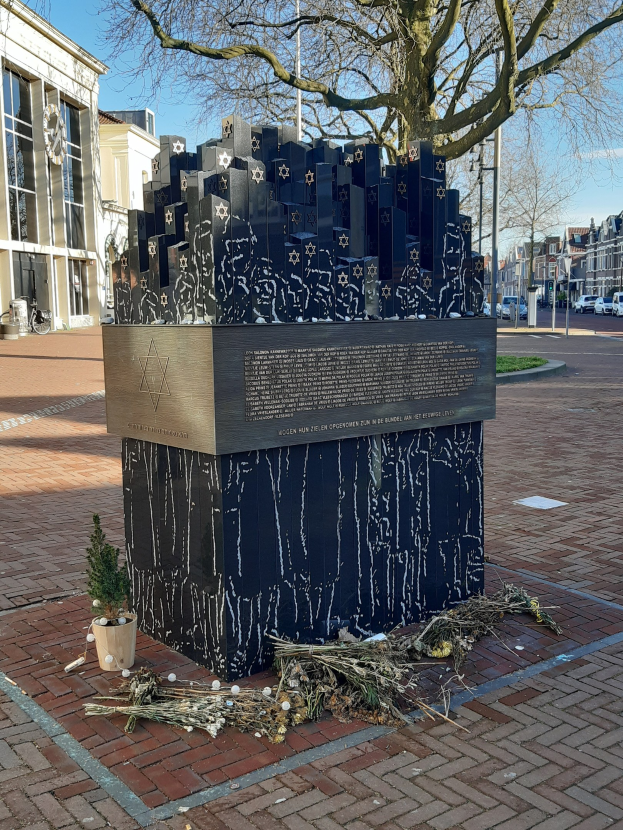 Stadtplatzgedächtnis für Holocaust-Opfer mit Baum vorne, Topfpflanze am Boden, verstreute trockene Blätter und umgebende Gebäude, Fahrzeuge, Mäste und Bäume.