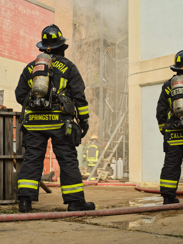 Zwei Feuerwehrleute in Schutzausrüstung stehen vor einem raucherfüllten Gebäude mit einer Leitung auf dem Boden, während andere Gebäude und Bäume im Hintergrund zu sehen sind.