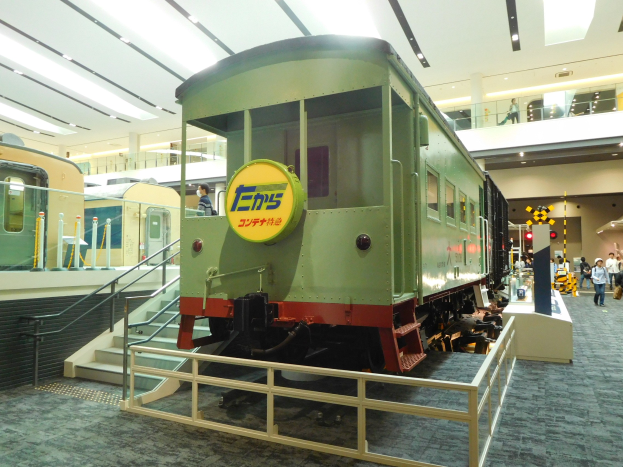 Ein grüner Zug in einem Museum mit Geländern und Treppen, umgeben von Menschen, einige halten Taschen, unter beleuchteter Decke.