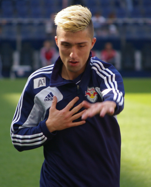 Ein junger Mann in einem blauen T-Shirt steht auf einem Fußballfeld und hat die Arme ausgestreckt, er sieht entschlossen aus und ist als Flügelstürmer zu erkennen.
