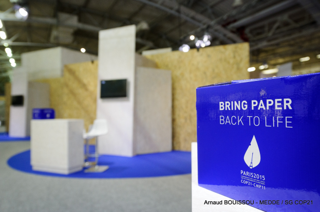 Messestand mit einem blauen und weißen "Bring Paper Back to Life"-Schild auf der rechten Seite, das eine unscharfe Sicht auf Stühle, Tische, einen wandmontierten Fernseher und Deckenleuchten im Hintergrund zeigt.