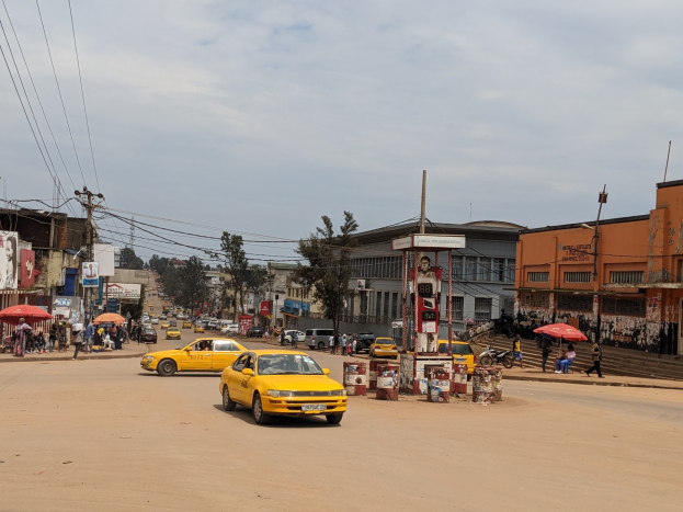Ein gelbes Taxi fährt auf einer belebten Stadtstraße mit Gebäuden an beiden Seiten. Fußgänger mit Regenschirmen gehen auf dem Gehweg, während sich der Himmel bewölkt.