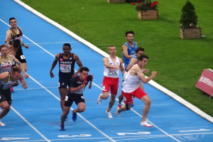 Eine Gruppe von Männern, die auf einer blauen Bahn laufen, umgeben von grünem Gras und Blumentöpfen mit einer Texttafel auf der rechten Seite, die auf einen Wettbewerb hinweist, wahrscheinlich die Leichtathletik-Europameisterschaften.