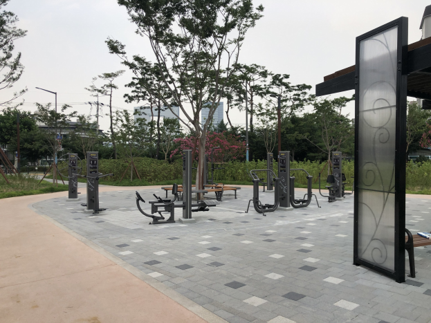 Outdoor park with fitness equipment, benches, trees, plants, grass, poles, lights, wires, and buildings against a sky backdrop.
