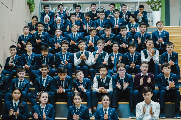 Eine Gruppe junger Menschen, wahrscheinlich ein Schulchor, sitzt auf einer Treppe und klatscht feierlich, mit Bücherregalen und Pflanzen im Hintergrund.